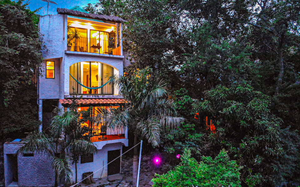 Fachada iluminada de las habitaciones del Jardín Secreto rodeada de árboles y vegetación al atardecer