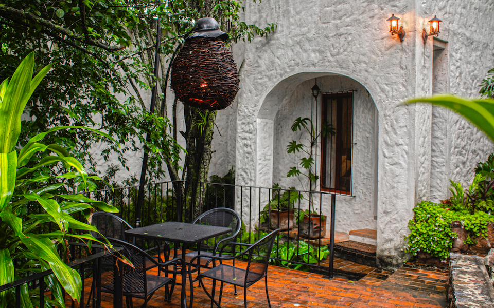 Terraza con mesa y sillas rodeada de vegetación, junto a fachada blanca con lámparas encendidas