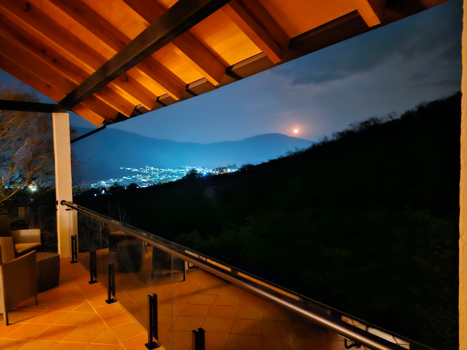 Vista nocturna desde la terraza de la Cabaña Las Águilas con luces de la ciudad y luna al fondo.