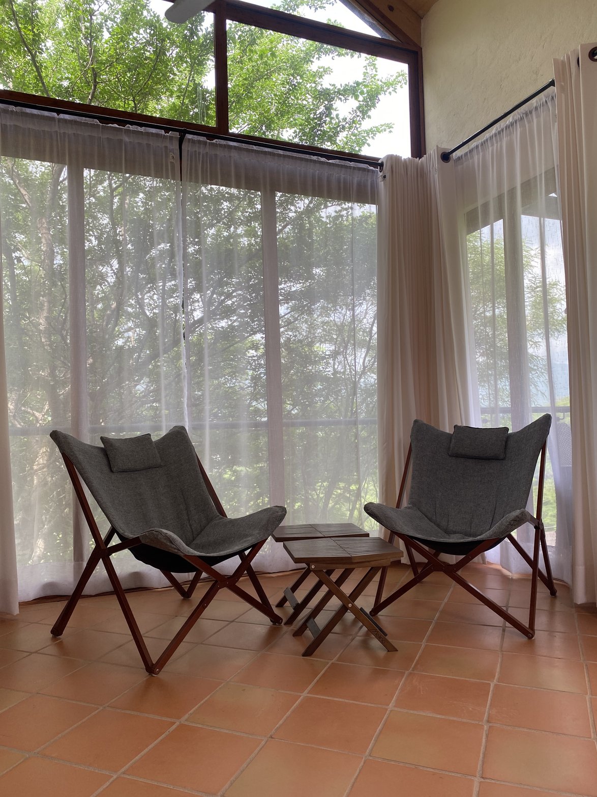 Rincón con sillas frente a grandes ventanales con cortinas y árboles al fondo.