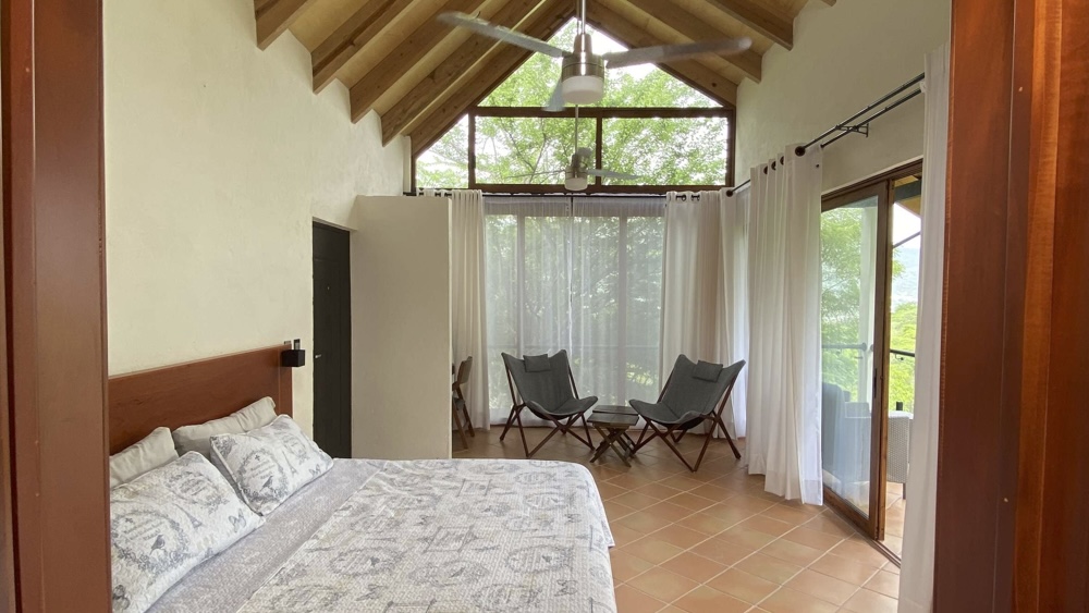 Interior de la Cabaña Las Águilas con cama king size, techo de madera a dos aguas, ventanales con cortinas blancas y vista panorámica al bosque