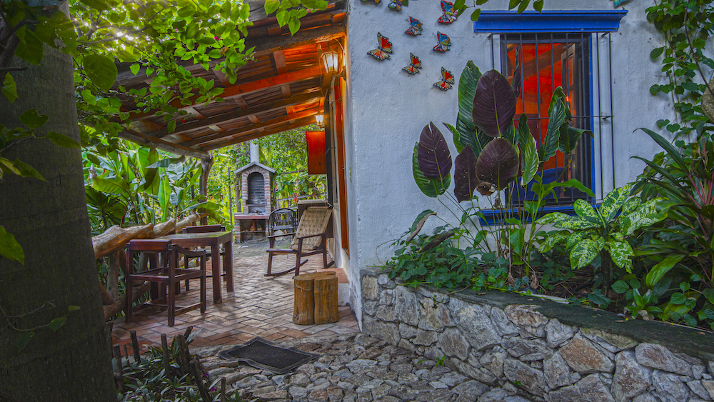 Porche acogedor de la Cabaña Mariposas con mesa, sillas y decoración de mariposas en la pared, rodeado de vegetación exuberante