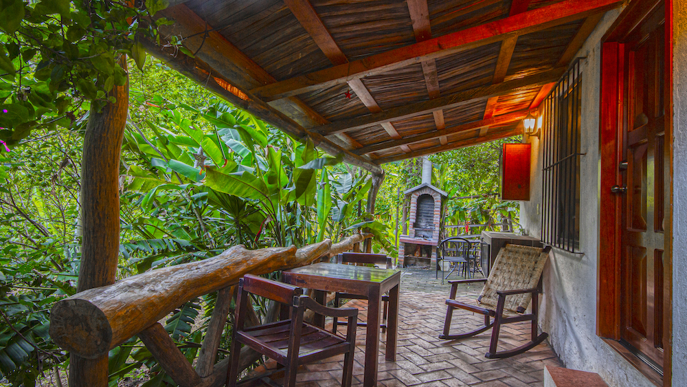 Terraza techada de la Cabaña Mariposas con sillas, mesa y mecedora, rodeada de plantas verdes y ambiente natural