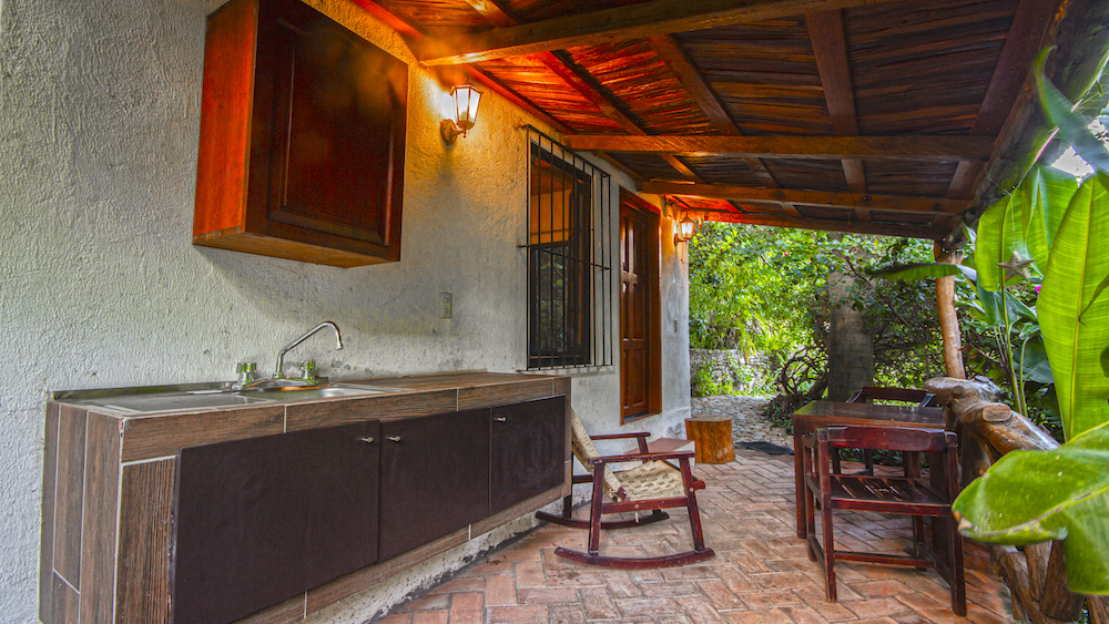Cocineta exterior de la Cabaña Mariposas con fregadero, mecedora y terraza techada rodeada de naturaleza