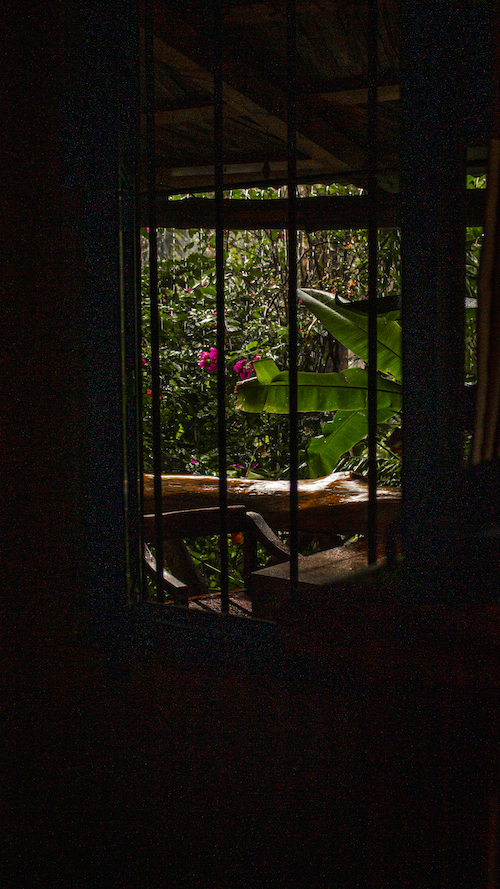 Vista desde el interior de la Cabaña Mariposas hacia el porche y el jardín, a través de una ventana con rejas