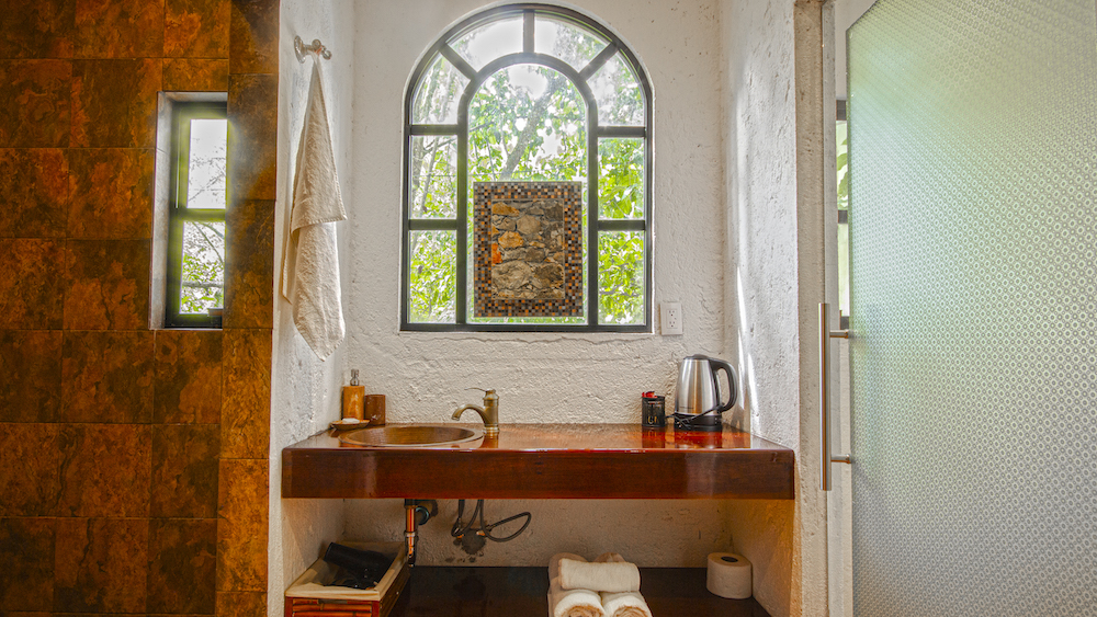 Pasillo de entrada a la Habitación El Cardenal con muro de piedra, puerta del baño y vista a la cama al fondo.