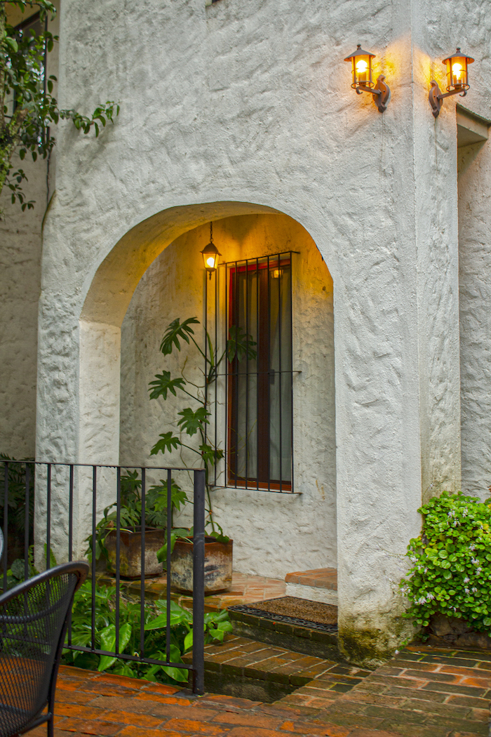 Entrada de la Habitación El Jardín Secreto con muros blancos texturizados, arco y faroles encendidos rodeada de vegetación.