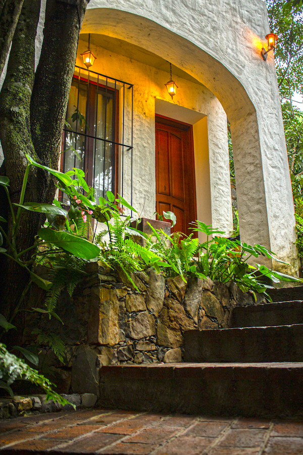 Escalinata hacia la entrada de la Habitación El Jardín Secreto, con puerta de madera, muros blancos y faroles iluminando entre plantas verdes.