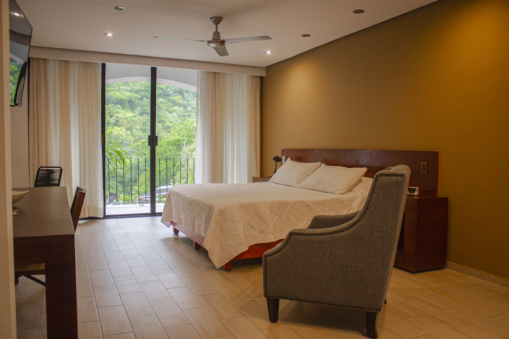 Interior de la Habitación El Jardín Secreto con cama king size, sillón gris, escritorio y gran ventanal con vista a la naturaleza.