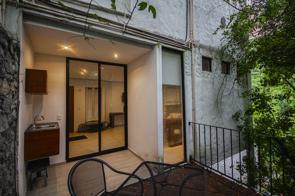 Terraza exterior de la Habitación El Jardín Secreto con mesa y sillas metálicas, tarja y ventanales de cristal.
