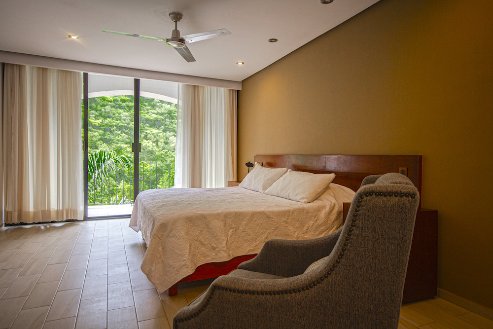 Vista de la Habitación El Jardín Secreto con cama king size, sillón gris y ventanal con vista a la vegetación.