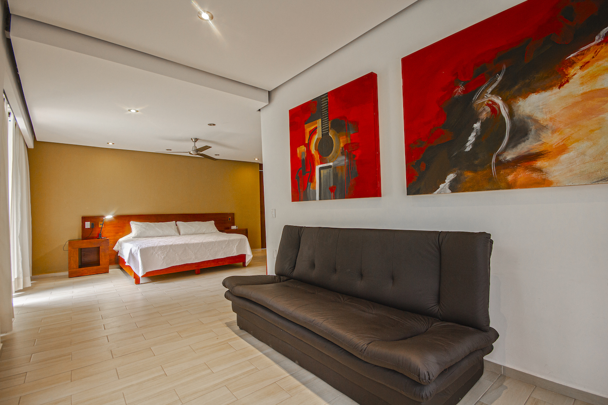 Vista interior de la Habitación El Jardín Secreto con cama king size al fondo y sofá frente a cuadros rojos decorativos.