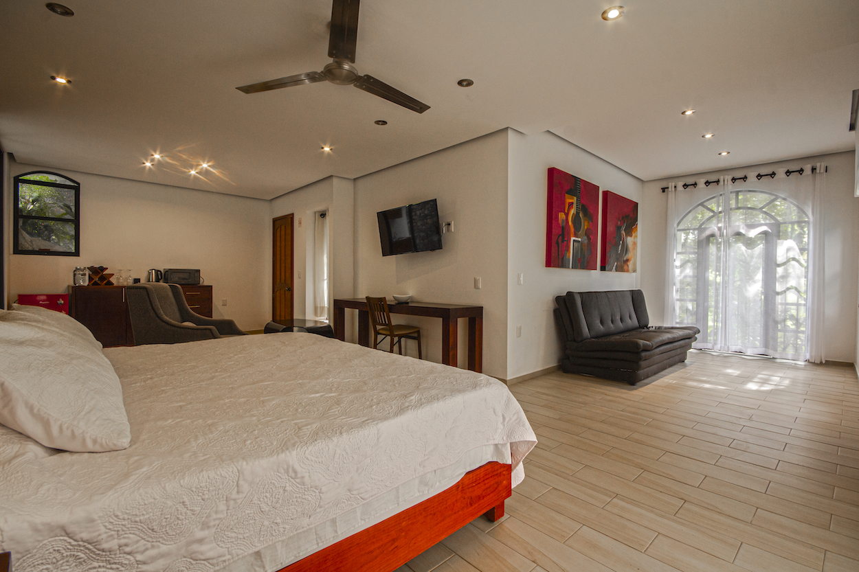 Vista panorámica de la recámara principal de la Habitación El Jardín Secreto con cama, sofá, escritorio y ventanales con luz natural.