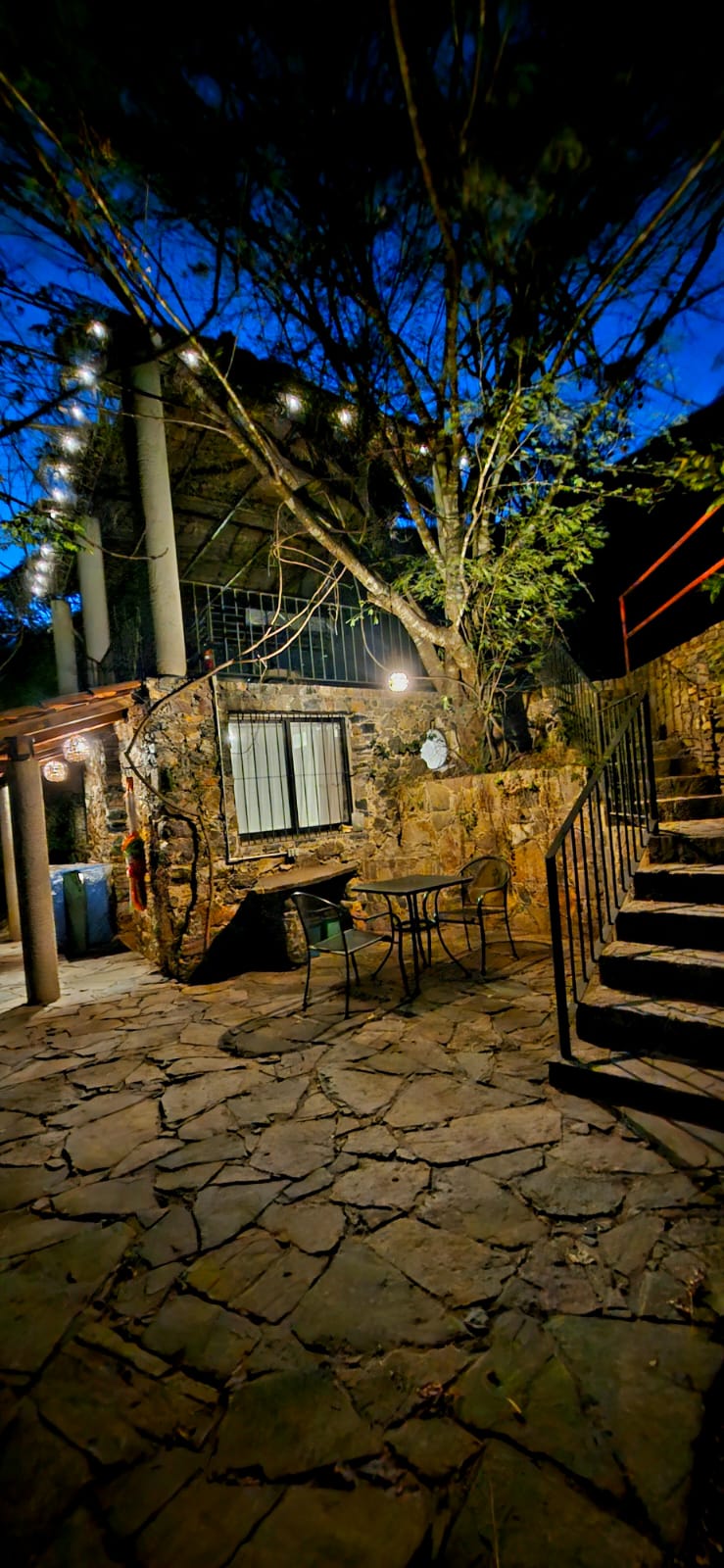 Vista nocturna de la terraza de la Habitación San Francisco con mesa y sillas metálicas sobre piso de piedra, escaleras y un árbol iluminado bajo el cielo azul profundo.