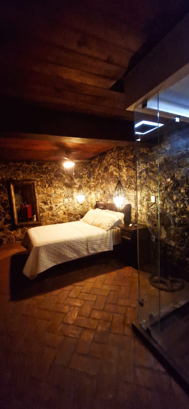 Habitación San Francisco con cama matrimonial iluminada por lámparas colgantes, muros de piedra y piso de madera, junto a regadera de cristal.