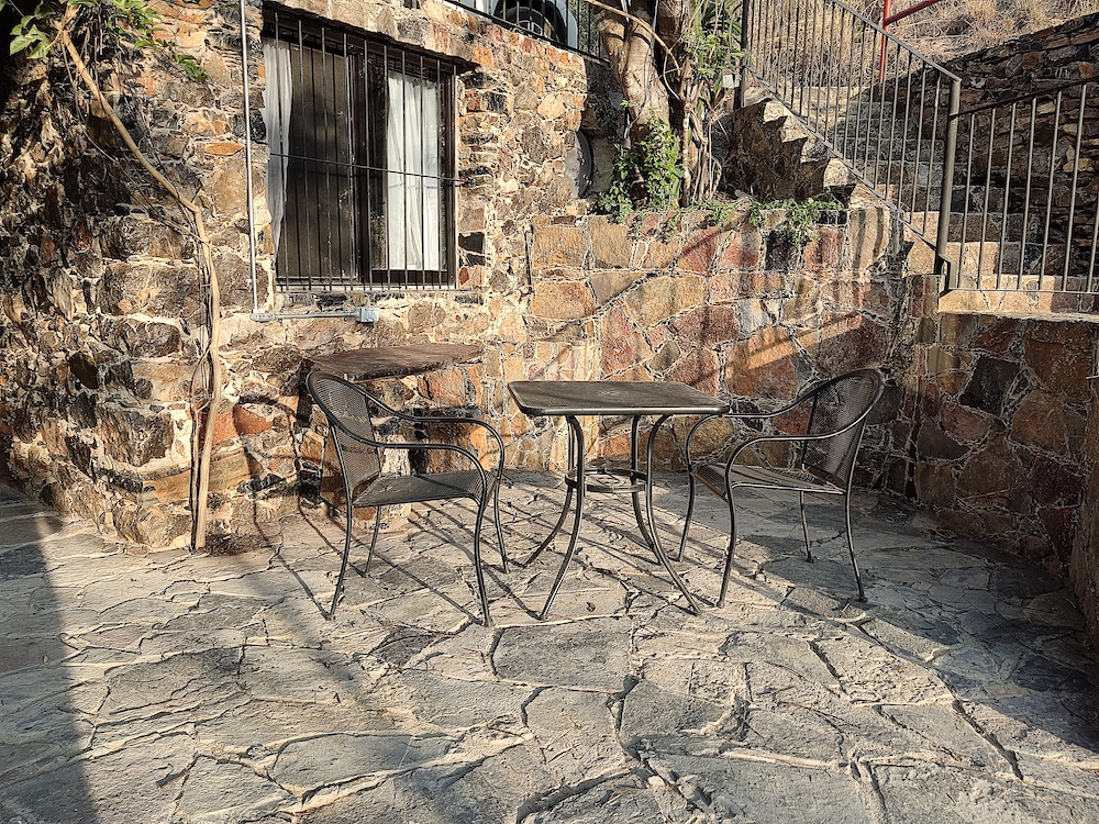 Terraza exterior de la Habitación San Francisco con mesa y tres sillas metálicas sobre piso de piedra, rodeada por muros de piedra y vegetación.