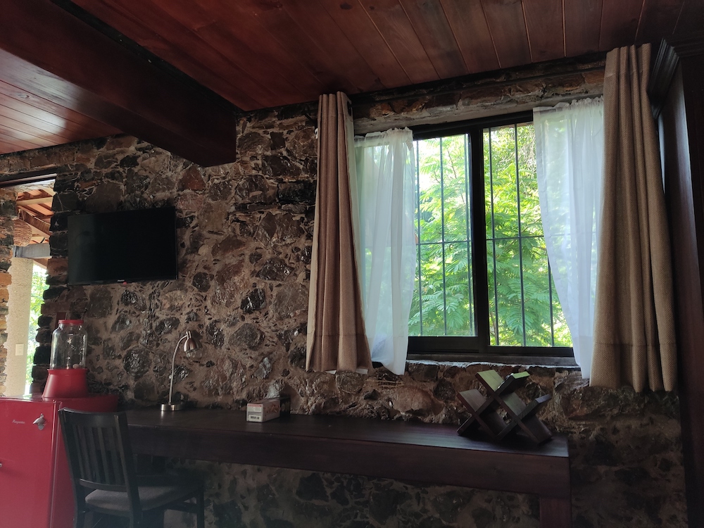 Interior de la Habitación San Francisco con escritorio de madera, televisor, refrigerador rojo y ventana con vista a los árboles.