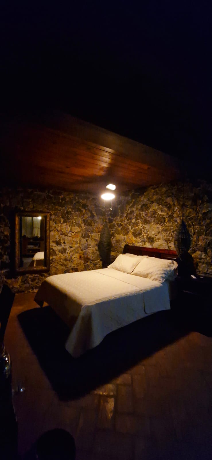 Habitación San Francisco de noche con cama matrimonial iluminada tenuemente, muros de piedra, techo de madera y espejo decorativo.