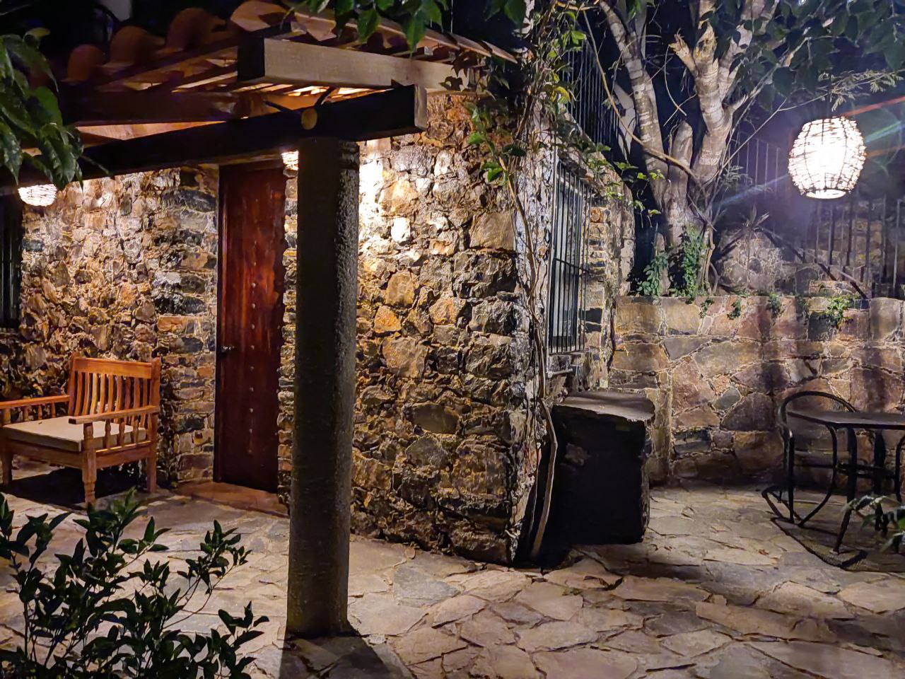 Entrada nocturna de la Habitación San Francisco con puerta de madera, banca de madera bajo un tejado y terraza de piedra iluminada con lámparas colgantes.