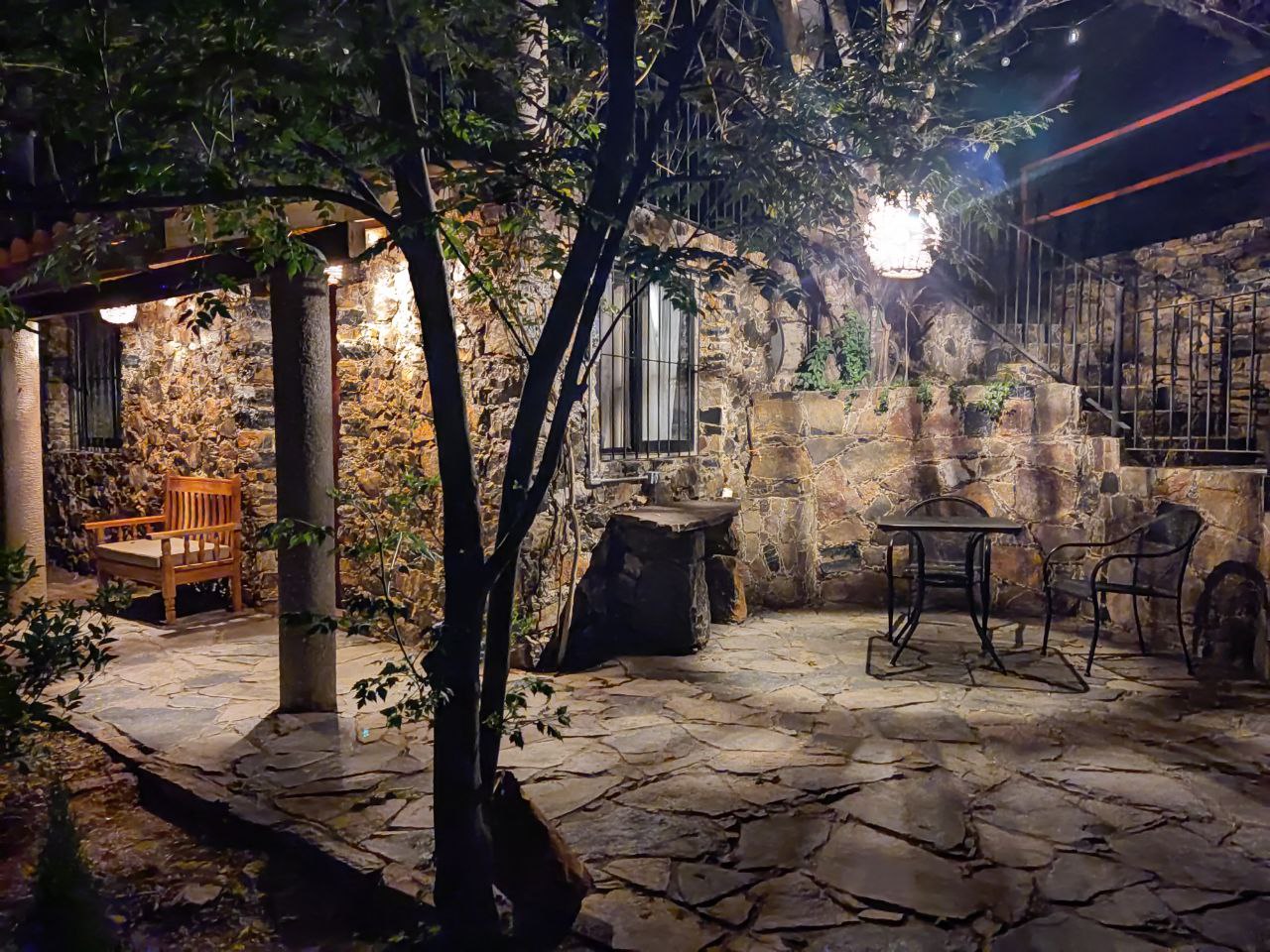 Vista nocturna del patio de la Habitación San Francisco con bancas, mesas y sillas sobre piso de piedra, iluminado entre árboles y faroles.