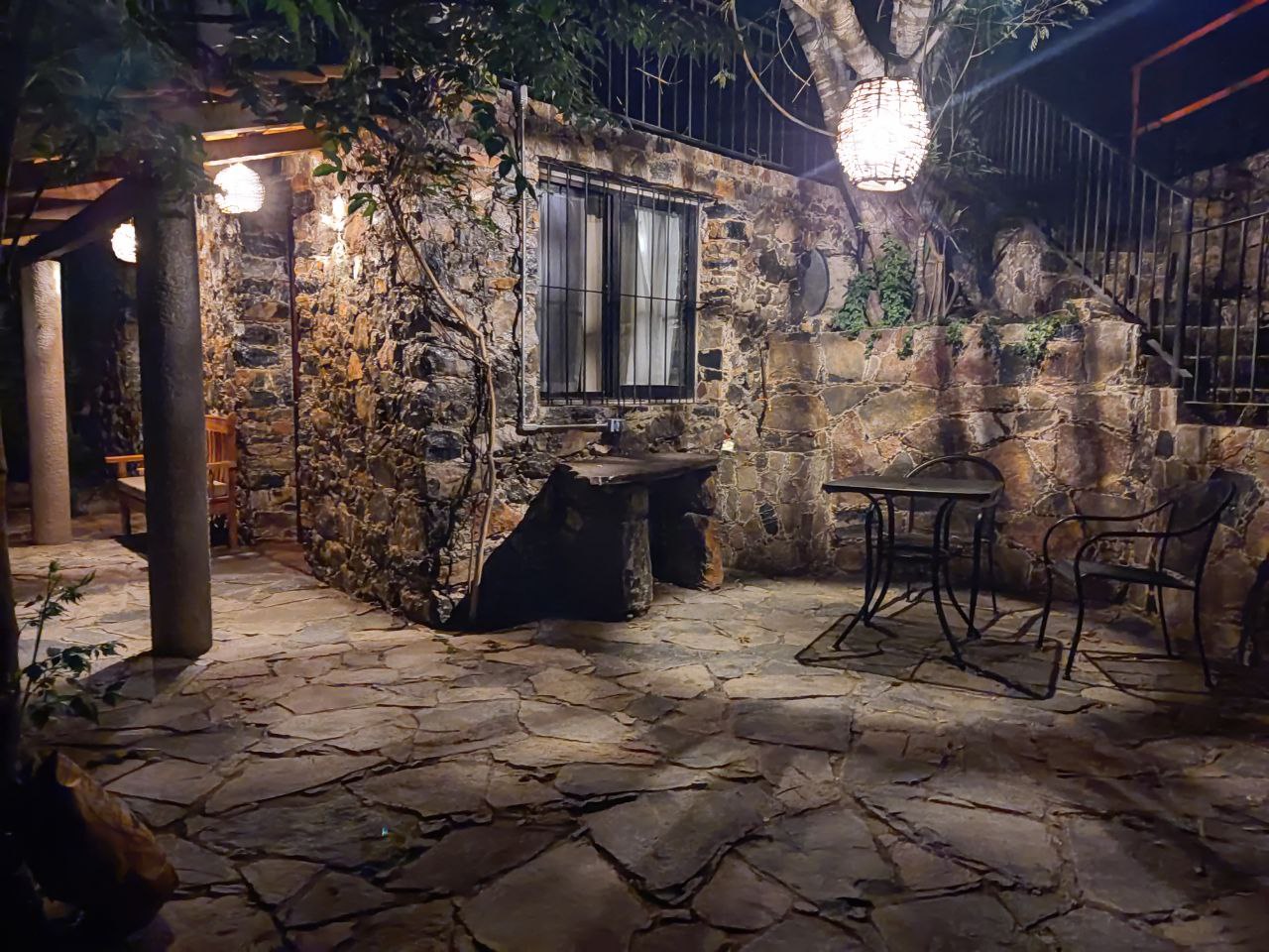 Patio nocturno de la Habitación San Francisco con mesa, sillas, banca de madera y una lámpara colgante entre los árboles, rodeado de muros de piedra.